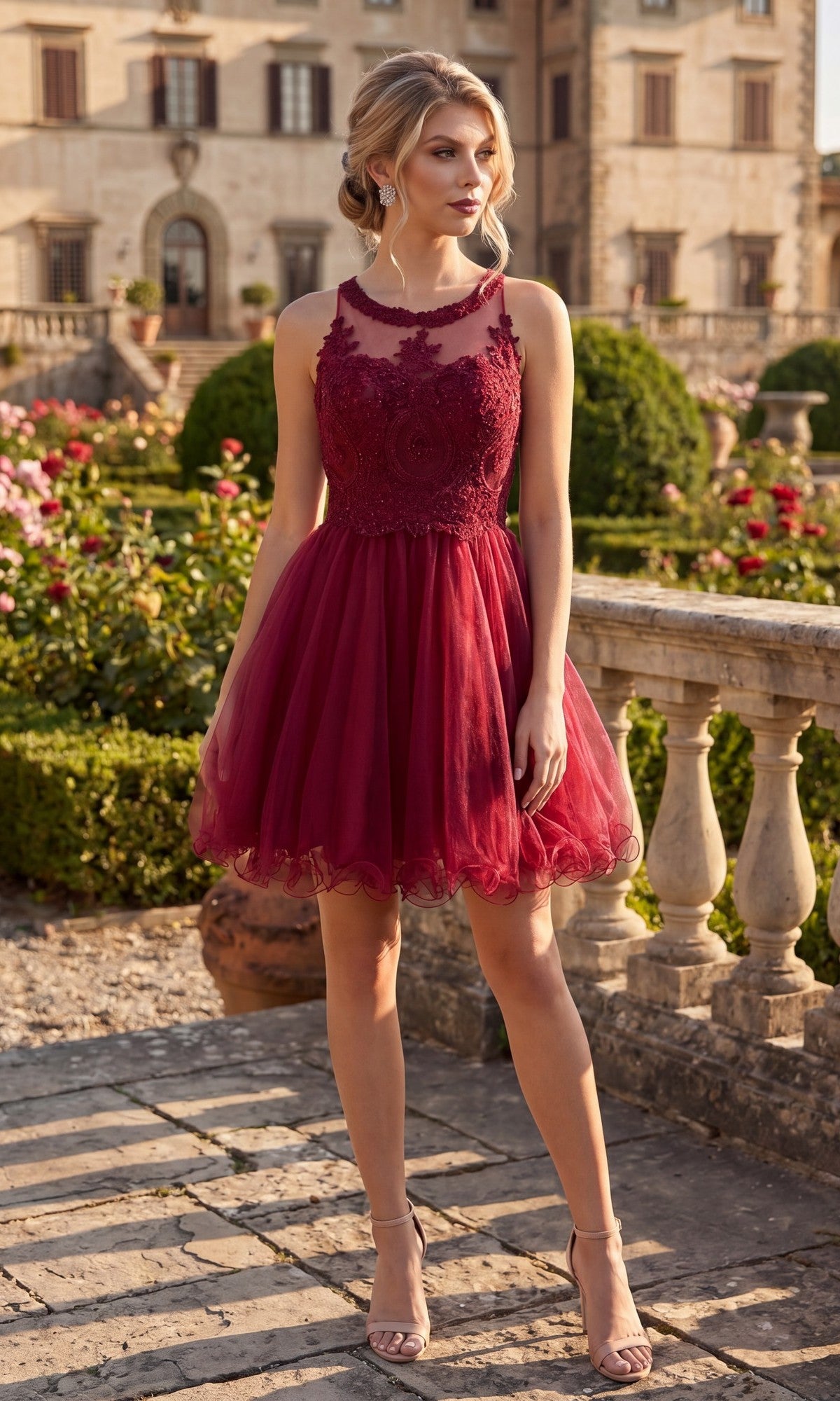 A woman wearing the Chicas DM4002 Short Homecoming Dress in burgundy, featuring a sleeveless fit-and-flare silhouette with an embroidered bodice, stands on a stone patio in a garden surrounded by trimmed bushes, flowers, and a historic building in the background.